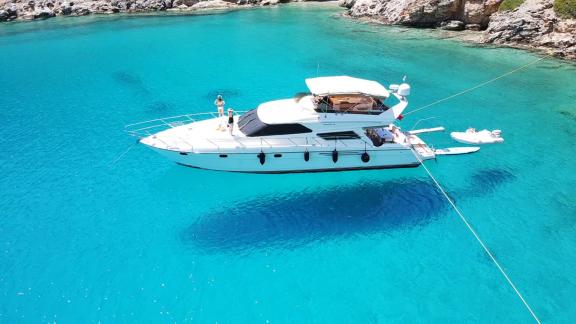 Motor yacht Hadron is anchored in clear turquoise waters with a rocky coastline in the background.